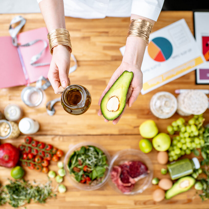 Dietitian holding avocado and olive oil above the table full of healthy food Dietitian holding avocado and olive oil above the table full of various healthy products, ketogenic diet concept